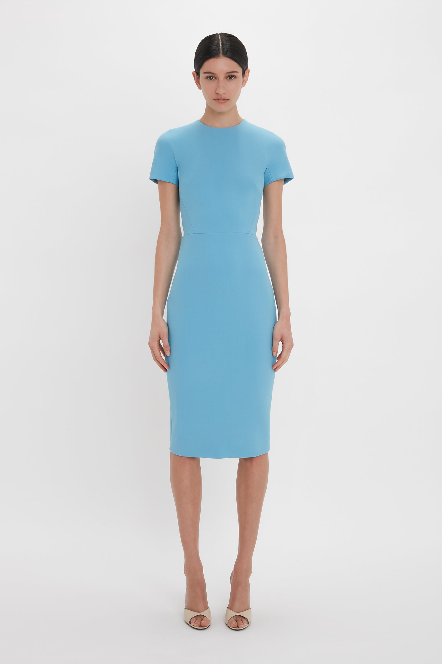 A person stands against a plain white background, wearing the Victoria Beckham Exclusive Fitted T-Shirt Dress In Periwinkle Blue and beige heels.