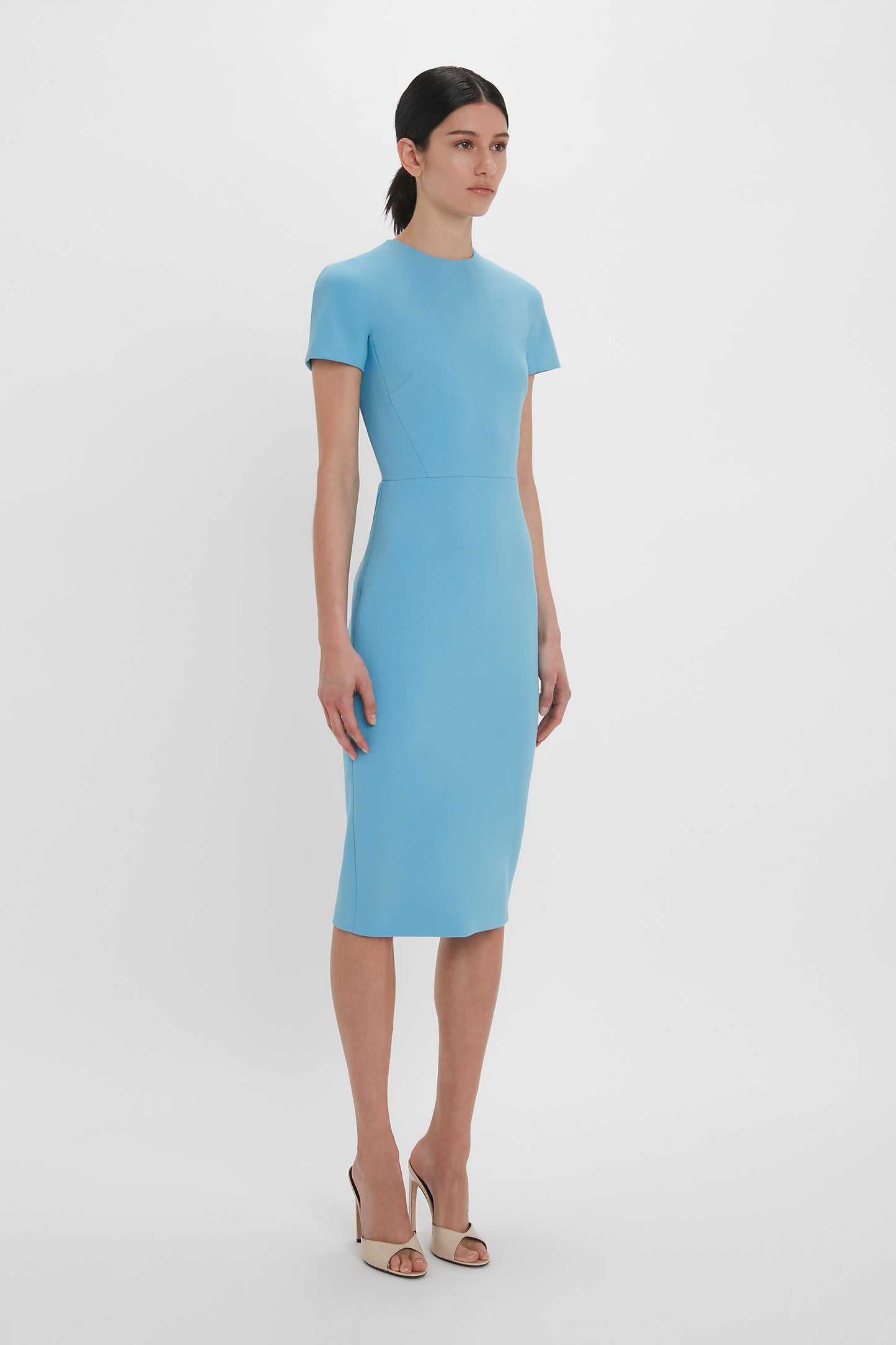 A woman stands against a plain background, wearing the Victoria Beckham Exclusive Fitted T-Shirt Dress In Periwinkle Blue with short sleeves and beige open-toe heels. Her hair is tied back in an elegant style, reminiscent of Victoria Beckham's iconic fashion sense.