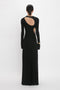 A person wearing a Victoria Beckham Cut-Out Jersey Floor-Length Dress In Black stands facing away from the camera. The elegant black evening gown features long sleeves and an open back, adding a touch of sophistication to its cutout designs.