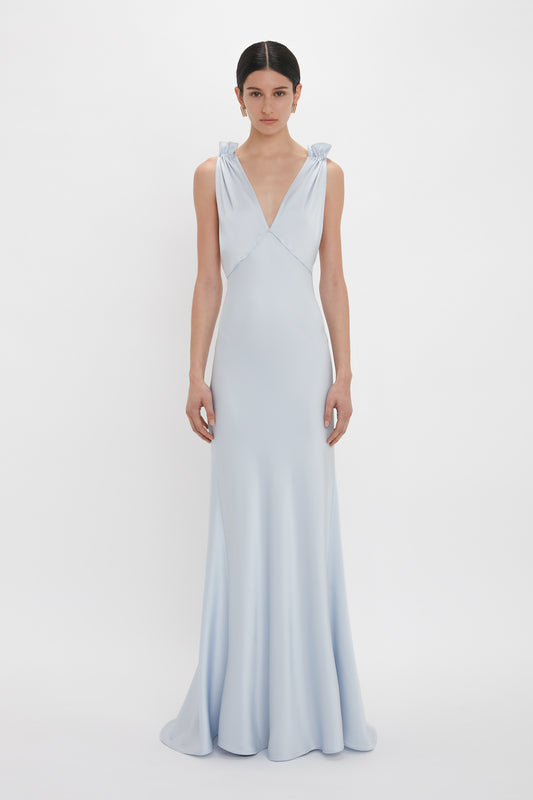 A person stands facing forward, wearing an Exclusive Gathered Shoulder Cami Floor-Length Gown In Ice Blue by Victoria Beckham made of luxurious crepe back satin with a deep V-neckline, against a plain white background.