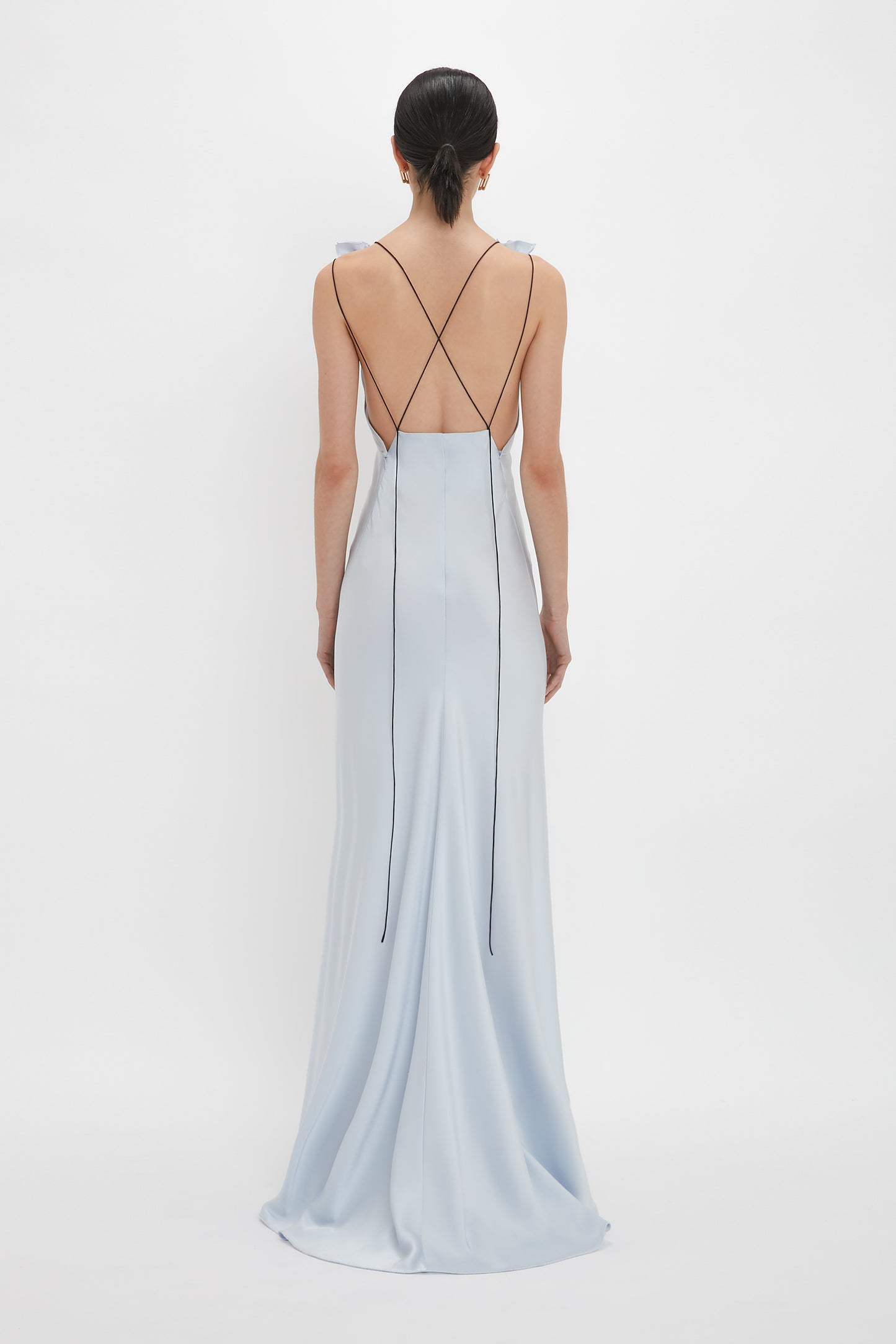 A woman in a light blue Exclusive Gathered Shoulder Cami Floor-Length Gown In Ice Blue by Victoria Beckham with an open-back and thin black straps crisscrossing at the back, stands facing away against a white background.