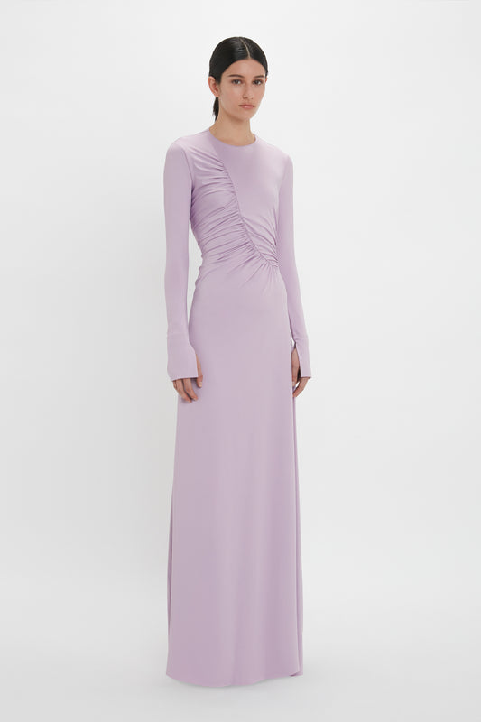 A person is wearing a long-sleeved, floor-length Ruched Detail Floor-Length Gown In Petunia by Victoria Beckham with ruched detailing on the front, standing against a plain white background.