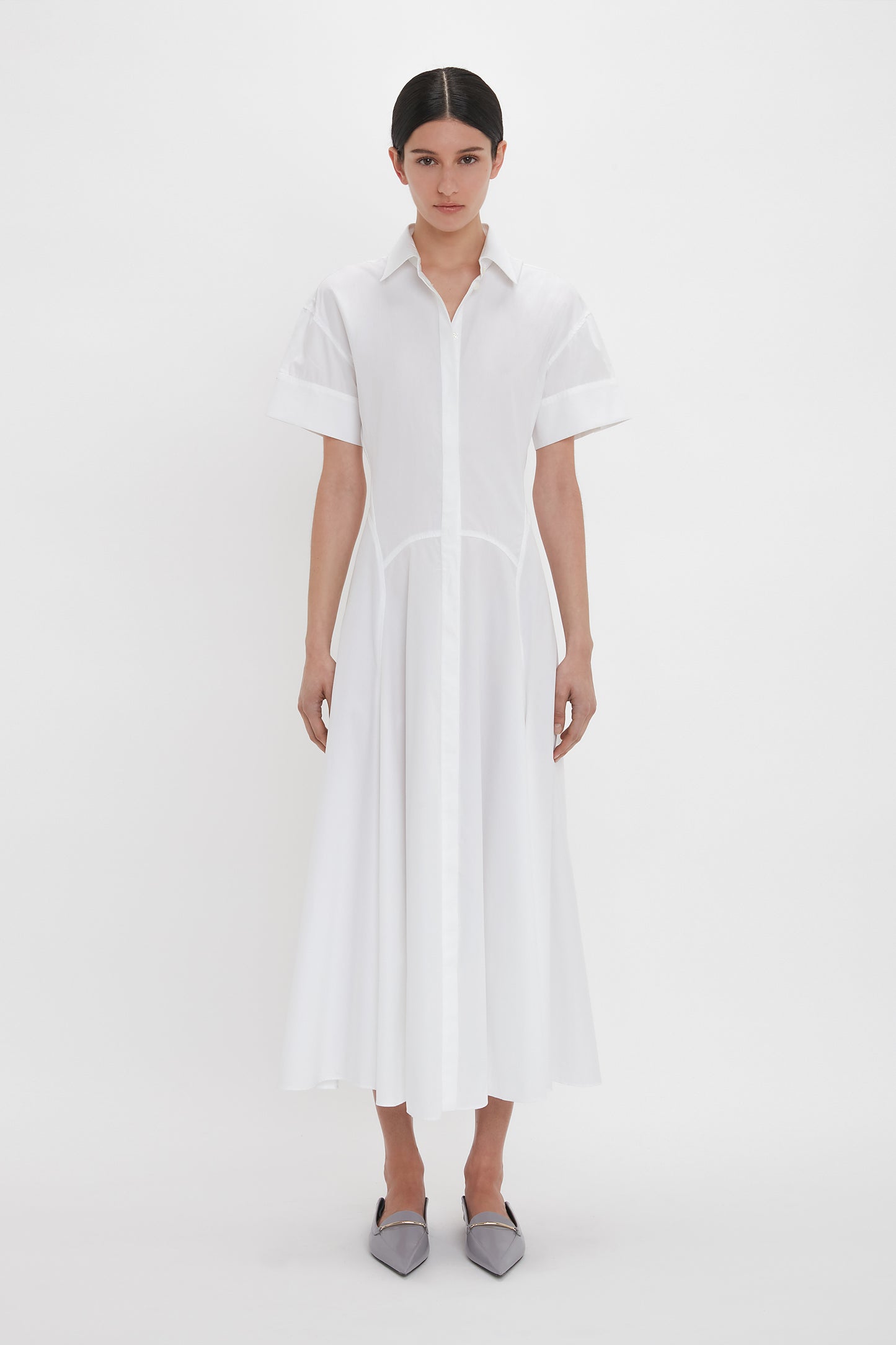 A person is wearing a Panelled Shirt Dress In White by Victoria Beckham that extends to mid-calf length with gold button cuffs and grey pointed flat shoes, standing against a plain white background.