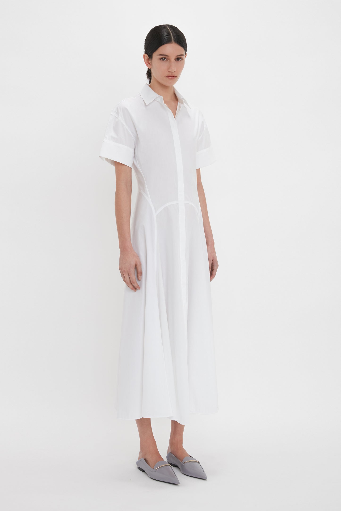 Person standing against a plain backdrop, wearing the Victoria Beckham Panelled Shirt Dress In White made from organic cotton poplin and grey pointed flats.