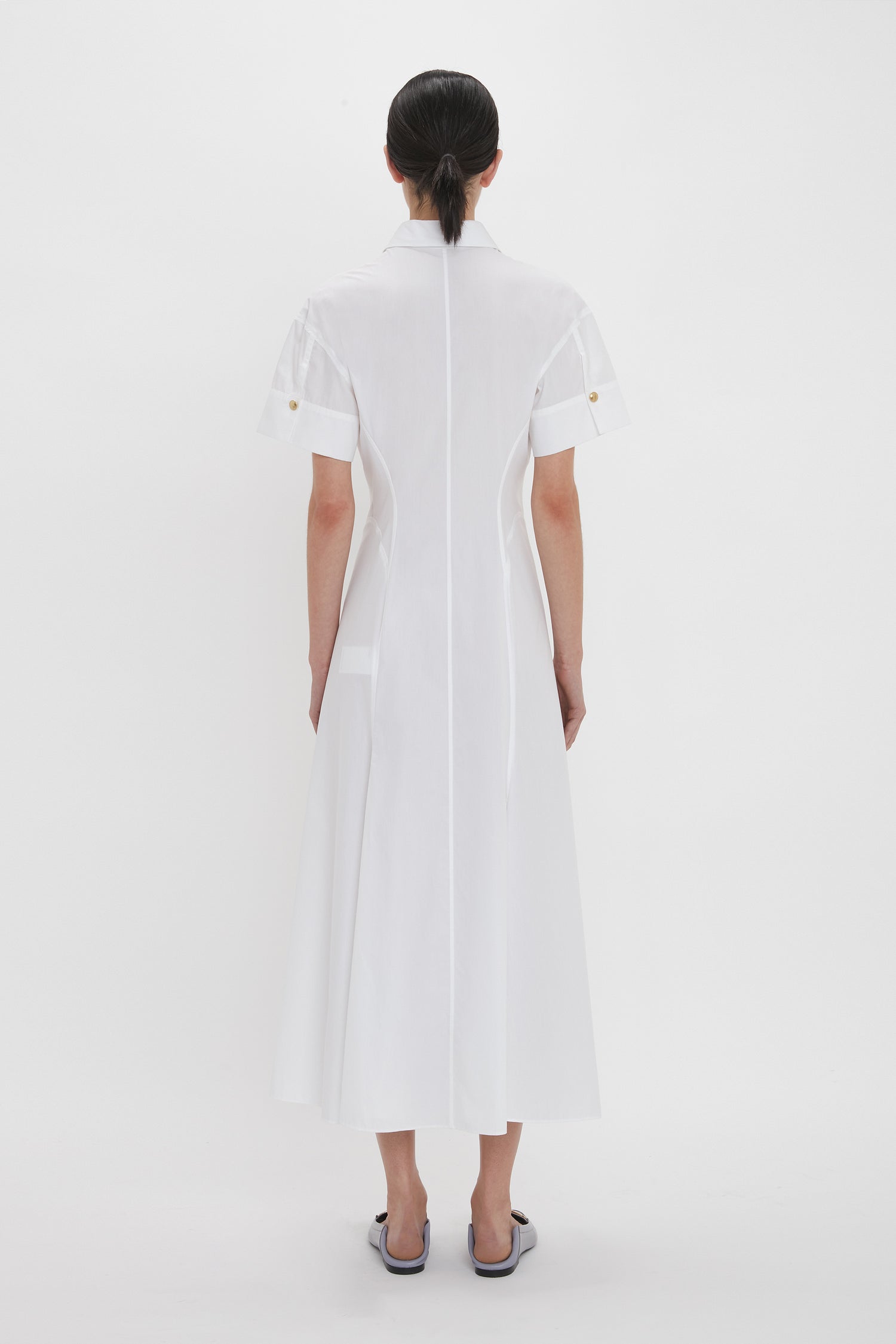 A person with dark hair stands facing away, wearing the Panelled Shirt Dress In White by Victoria Beckham, made of organic cotton poplin. The ankle-length dress features gold button cuffs near the sleeves. White background.