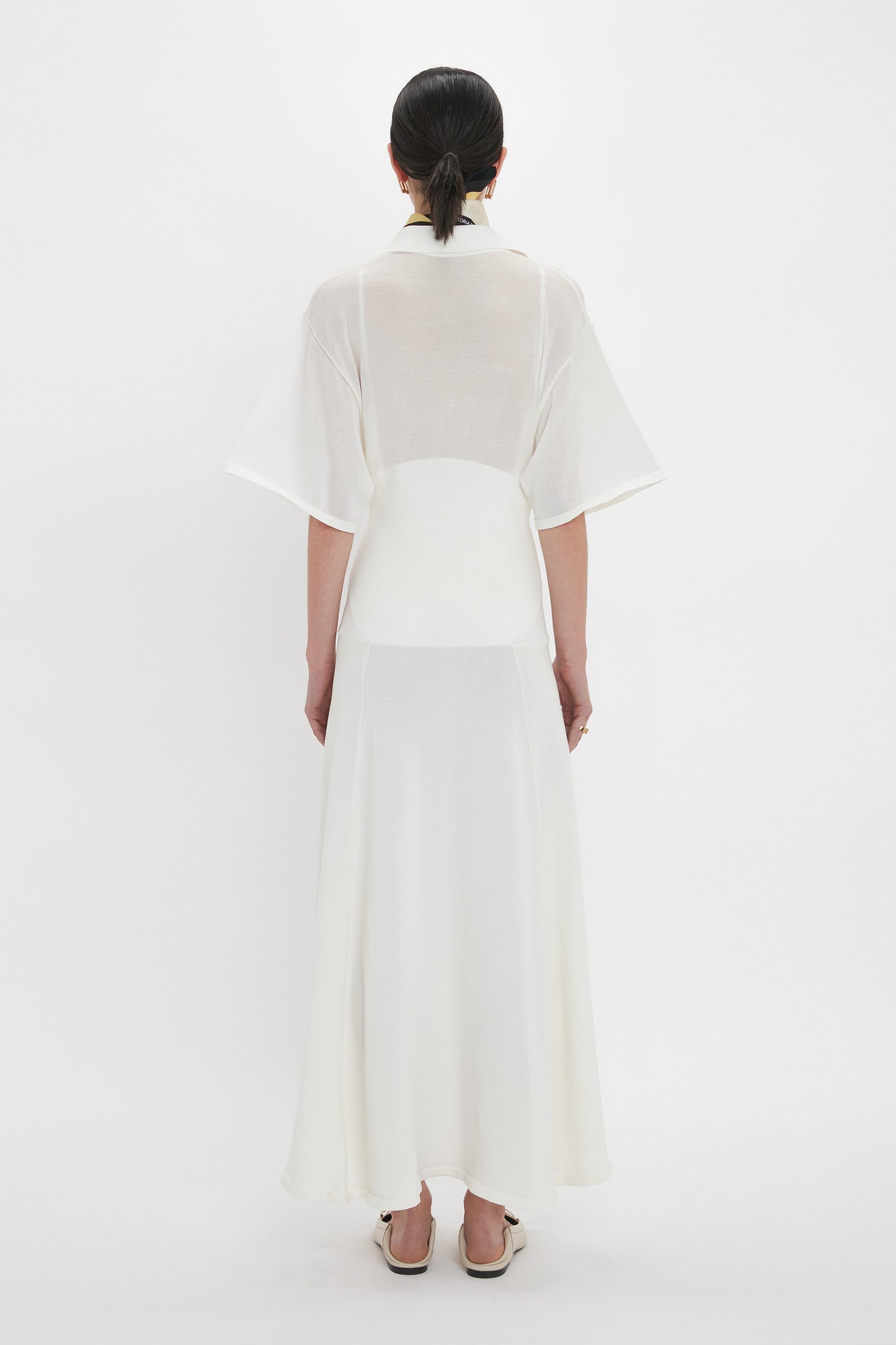 A person with dark hair in a bun is standing with their back to the camera, wearing a long, white, semi-sheer Panelled Knit Dress In White from Victoria Beckham with short sleeves. The background is plain white.