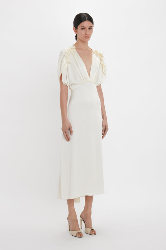 A woman stands wearing a cream-colored, mid-length dress with a deep V-neck and short, gathered sleeves. She has her hair pulled back and is wearing Victoria Beckham Classic Mule In Macadamia calf Leather with a seductive curved heel.