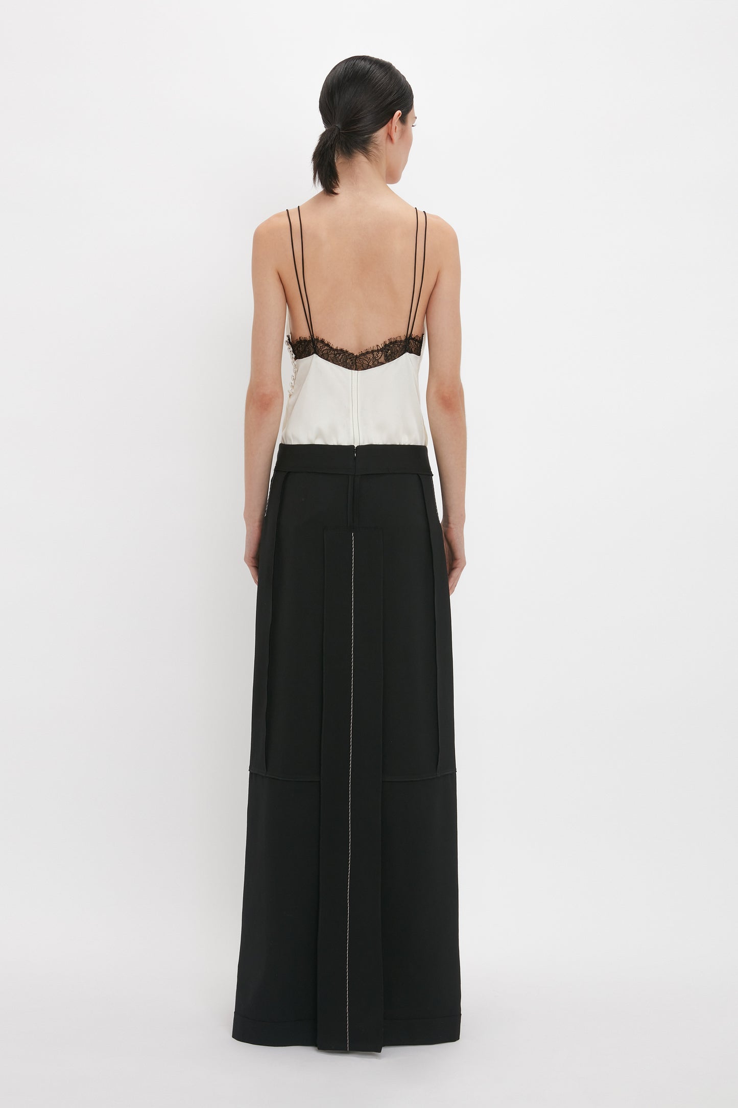 A person with dark hair, wearing a Lace Detail Cami Top In Harvest Ivory by Victoria Beckham and a long black skirt, stands facing away from the camera against a plain white background.