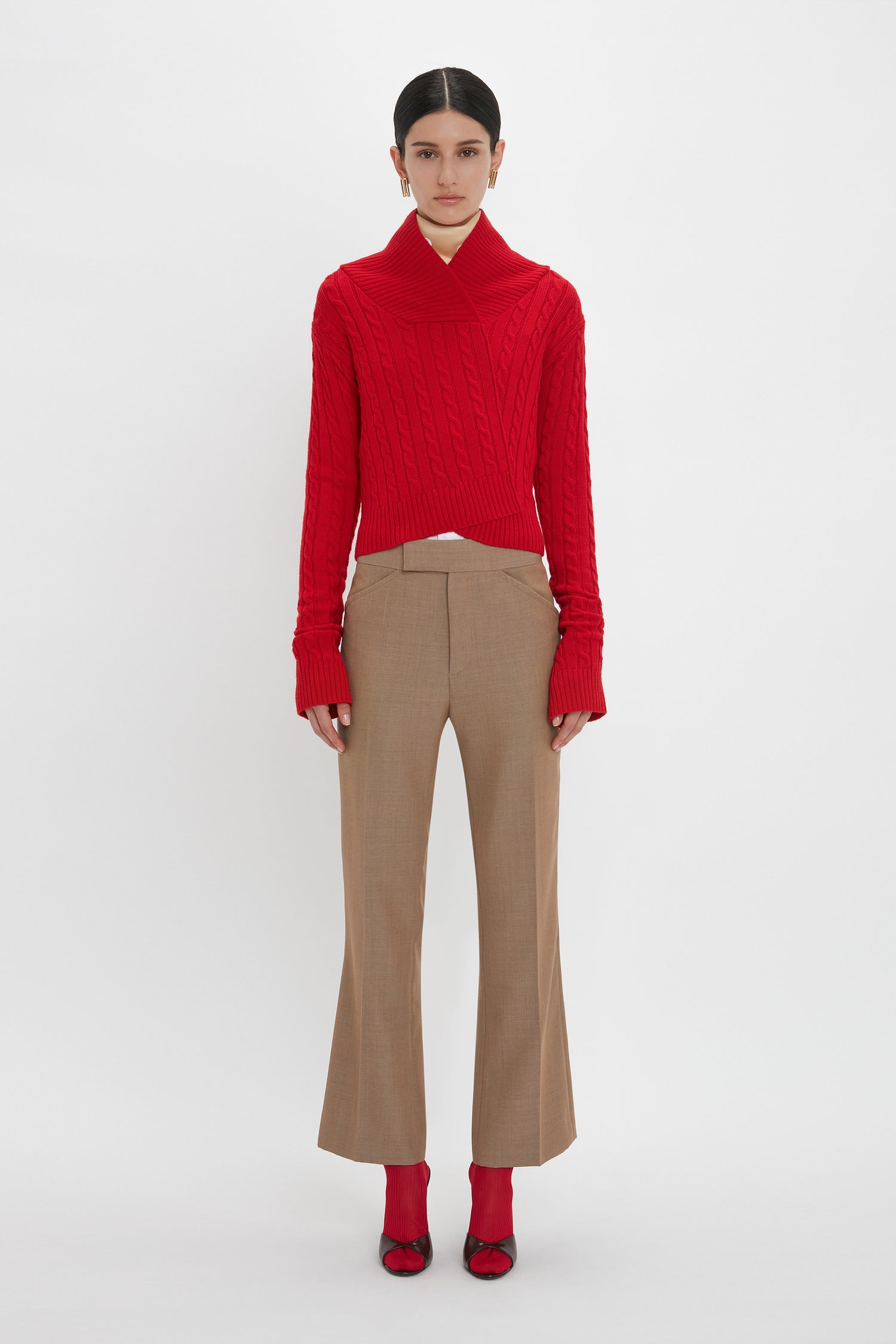 A person stands against a white background, clad in luxury knitwear. They're wearing the Wrap Detail Jumper In Red by Victoria Beckham, beige trousers, and red shoes. Their hair is tied back as they look straight ahead.