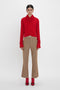 Person wearing a red knit sweater, Victoria Beckham's Wide Cropped Flare Trouser In Tobacco, and red high-heeled shoes stands against a plain white background.