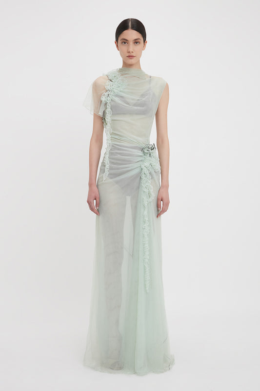A person stands wearing a sheer, pale green Gathered Tulle Detail Floor-Length Dress In Jade by Victoria Beckham with intricate floral detailing and a high neckline. The limited edition sea foam green tulle creation is set against a plain white background.