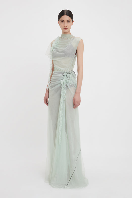 A person standing against a plain white background, wearing a sea foam green tulle gown with floral embellishments and a cinched waist. This limited edition Gathered Tulle Detail Floor-Length Dress In Jade by Victoria Beckham features exquisite detailing and elegant sheer fabric.