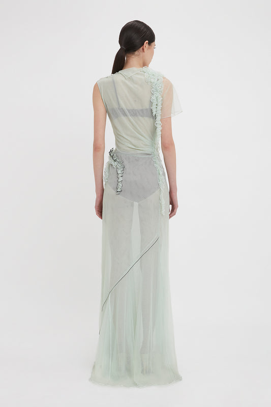 A person with long dark hair stands facing away, wearing a limited edition, sheer sea foam green Victoria Beckham Gathered Tulle Detail Floor-Length Dress In Jade adorned with floral appliqués.