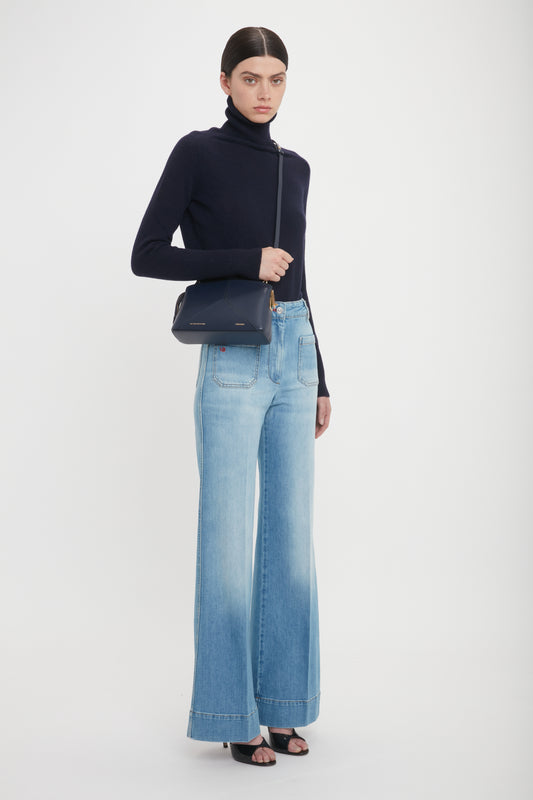 A person stands against a plain background, wearing a black turtleneck, light blue wide-leg jeans, and black heeled sandals, while holding an Exclusive Victoria Crossbody Bag In Navy Leather by Victoria Beckham.