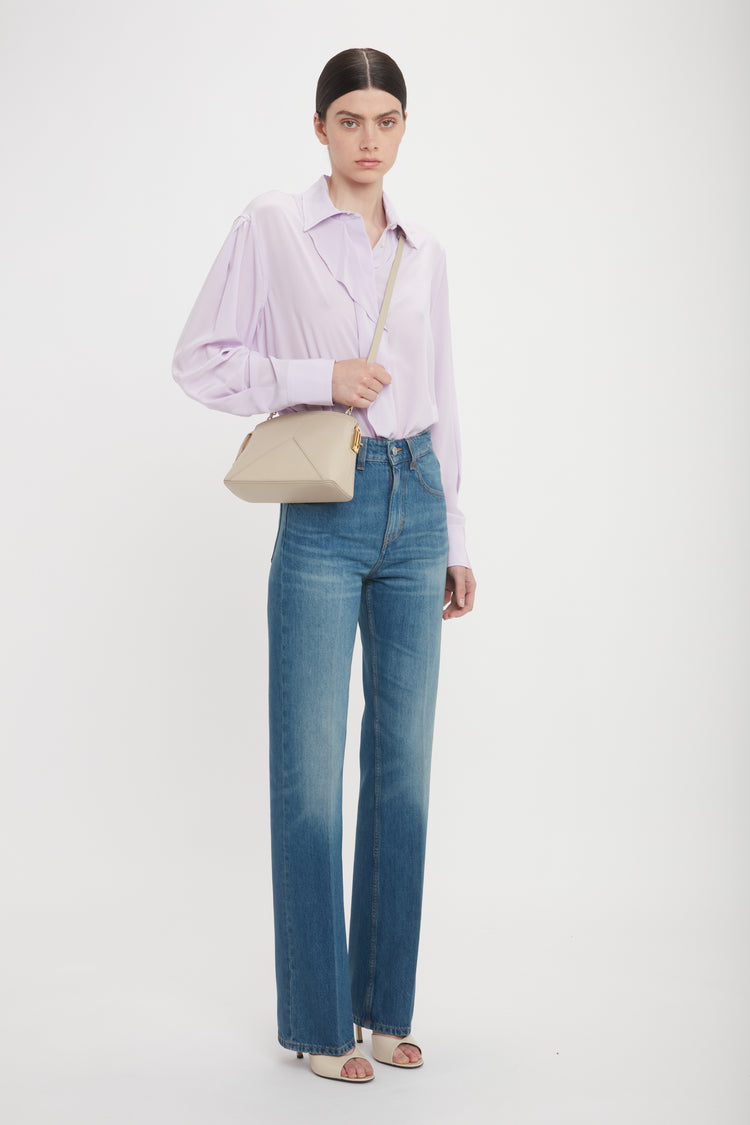 Person stands against a plain white background, wearing a light lavender shirt, high-waisted blue jeans, and open-toed heels. They carry a Victoria Beckham Victoria Crossbody Bag In Taupe Leather with structured design.