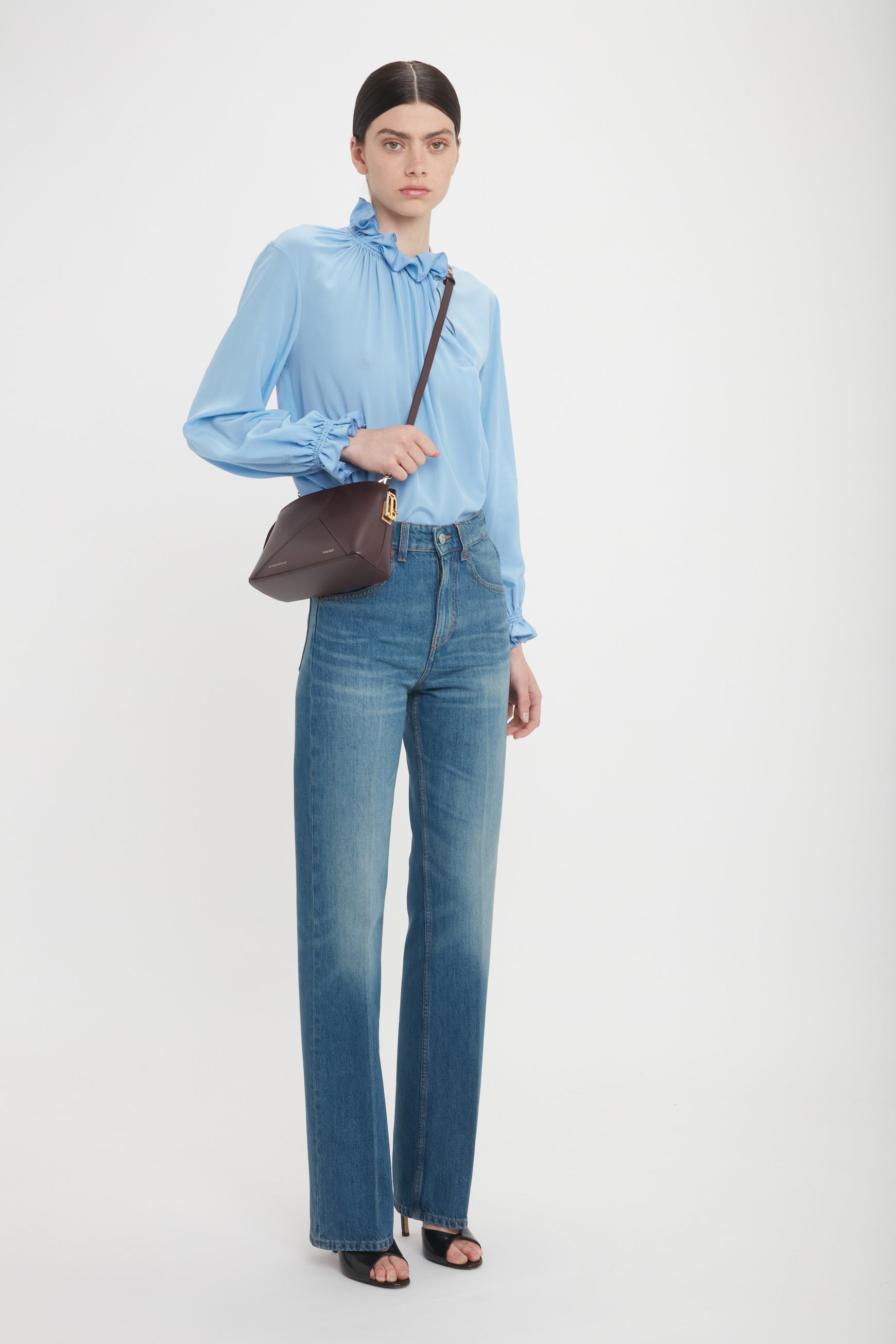 Person wearing a light blue blouse, high-waisted blue jeans, black open-toed heels, and carrying an Exclusive Victoria Crossbody Bag In Brown Leather by Victoria Beckham. The brown calf leather bag stands out as they pose against a plain white background.