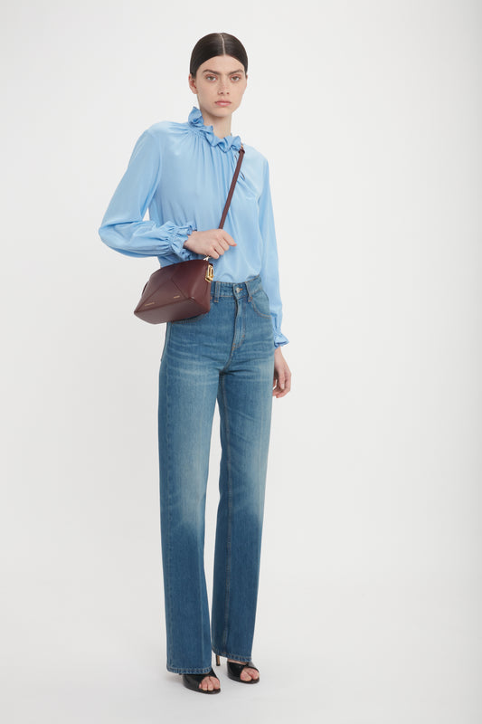 A person stands against a plain background wearing a light blue blouse, high-waisted jeans, open-toed black shoes, and a burgundy Victoria Crossbody Bag In Burgundy Leather by Victoria Beckham. The stylish bag features calf leather and a padlock closure. They have dark hair tied back.