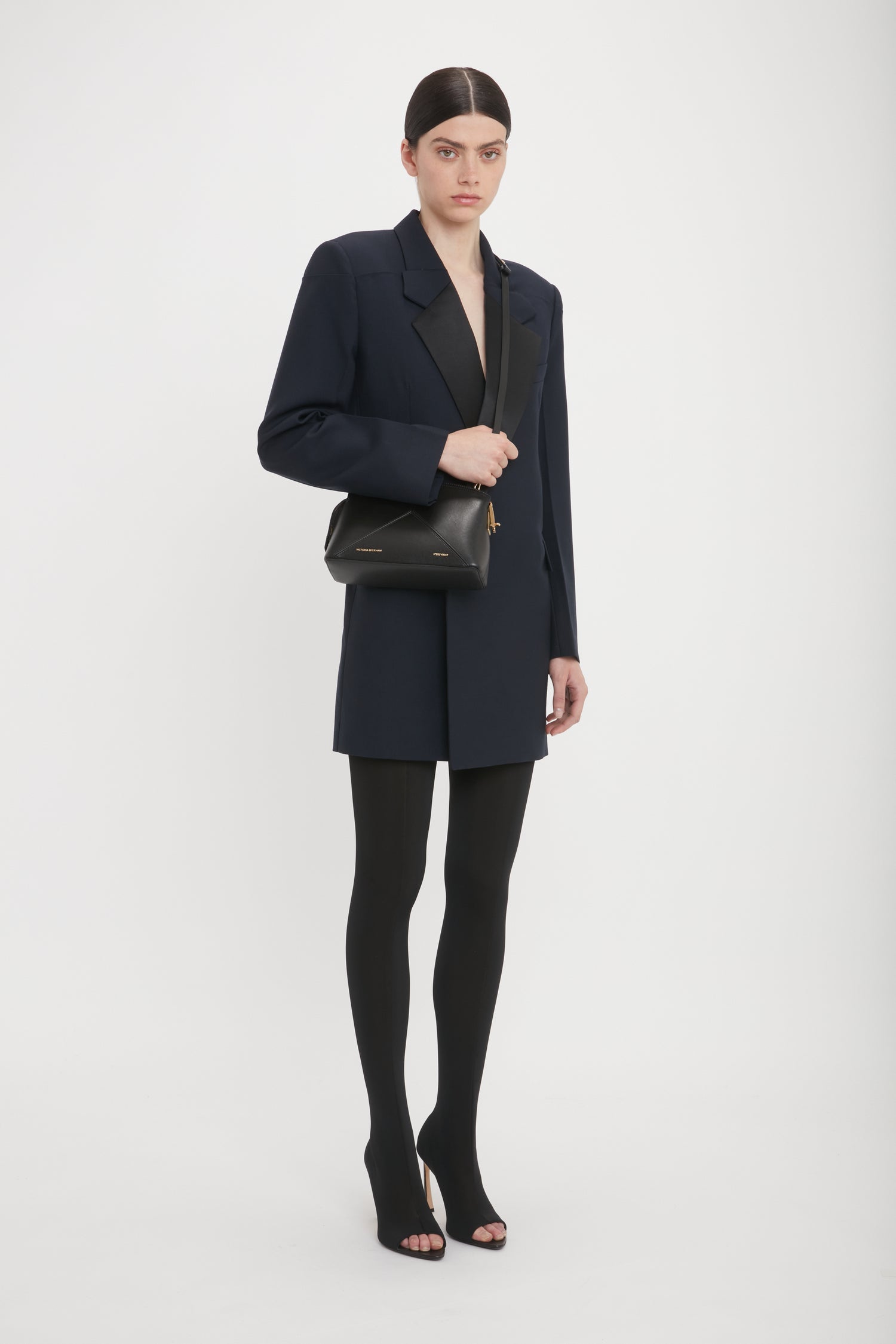 A person stands against a plain background, wearing a dark blazer and tights. They carry a Victoria Beckham Victoria Crossbody Bag In Black Leather with an adjustable strap, paired with open-toe high heels.