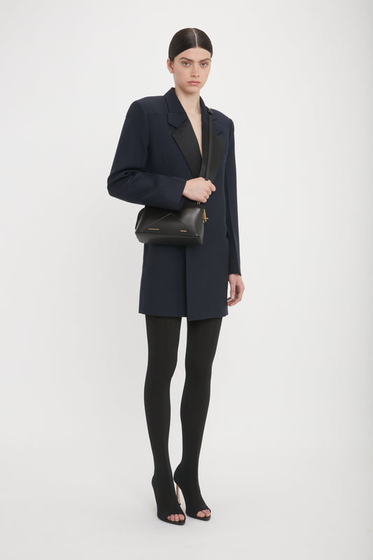 A person stands against a plain background, wearing a dark blazer and tights. They carry a Victoria Beckham Victoria Crossbody Bag In Black Leather with an adjustable strap, paired with open-toe high heels.