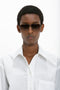 A person with short hair is wearing Victoria Beckham Double Bridge V-Line Navigator In Gold-Black-Brown sunglasses and a white button-up shirt with a pocket, standing against a white background.