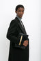 A person in a Double Breasted Tuxedo Coat in Black by Victoria Beckham with masculine tailoring and a gray turtleneck holds a black textured clutch with a gold accent, standing against a plain white background.