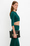 A woman with long brown hair is posing against a white background. She is wearing a teal VB Body Cropped Cardi In Lurex Green and matching skirt by Victoria Beckham that highlight her form-fitting silhouette, holding a black clutch with a gold chain accent.