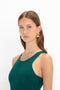 A woman with long brown hair wears a VB Body Sleeveless Dress In Lurex Green by Victoria Beckham and gold geometric earrings, posing against a white background, embodying the essence of a new-season wardrobe.