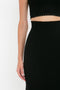 Cropped view of a person wearing a classic black sleeveless top and a VB Body Scallop Trim Flared Skirt In Black by Victoria Beckham, showing their left arm and hand against a plain white background.