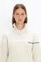 A person with long brown hair is wearing a cream-colored, **Collar Detail Jumper In Natural**, textured knit sweater with a high collar and a black stripe on the sleeve, standing against a plain white background, radiating casual sophistication. This stunning piece is by **Victoria Beckham**.