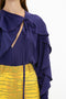 A person wearing a Tie Detail Ruffle Blouse In Ultraviolet by Victoria Beckham with a V neckline, perfectly paired with a high-waisted yellow patterned skirt.