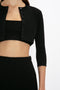 A woman is wearing a Victoria Beckham VB Body Cropped Cardigan In Black with buttons over a black top and a high-waisted black skirt. The image captures her upper body and waist, highlighting the form-fitting silhouette that emphasizes her feminine sensibility.