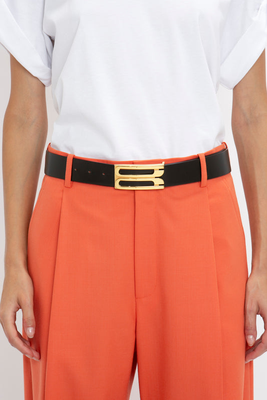 A person wearing an orange pair of pants with a Jumbo Frame Belt In Black Leather by Victoria Beckham featuring smooth calf leather and gold hardware, and a white shirt.