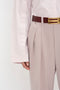 A close-up of a person wearing the Victoria Beckham Button Detail Cropped Shirt In Rose Quartz tucked into high-waisted beige trousers with a brown belt featuring a gold buckle. The person's left arm and hand are visible by their side, showcasing the button detail on the shirt's cuff.