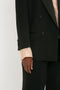 A person wearing a Victoria Beckham Satin Lapel Tuxedo Jacket in Black and pants with a light-colored shirt underneath. The image focuses on the lower half of the jacket, emphasizing the satin-covered buttons, and the person's hand.