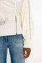 Person wearing a Victoria Beckham Asymmetric Gather Detail Top In Cream with blouson sleeves in a white, textured long-sleeve shirt with black detailing and blue jeans, visible from mid-torso to the hip area.