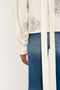 A person stands with their back to the camera, wearing Victoria Beckham's Asymmetric Gather Detail Top In Cream made from 100% silk and high-waisted blue denim jeans. The blouse features elegant blouson sleeves and two long fabric ties hanging down the back.