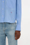 A person wearing a relaxed fit, light blue button-up **Cropped Long Sleeve Shirt In Oxford Blue by Victoria Beckham** and blue jeans. Only their lower torso and one arm are visible.