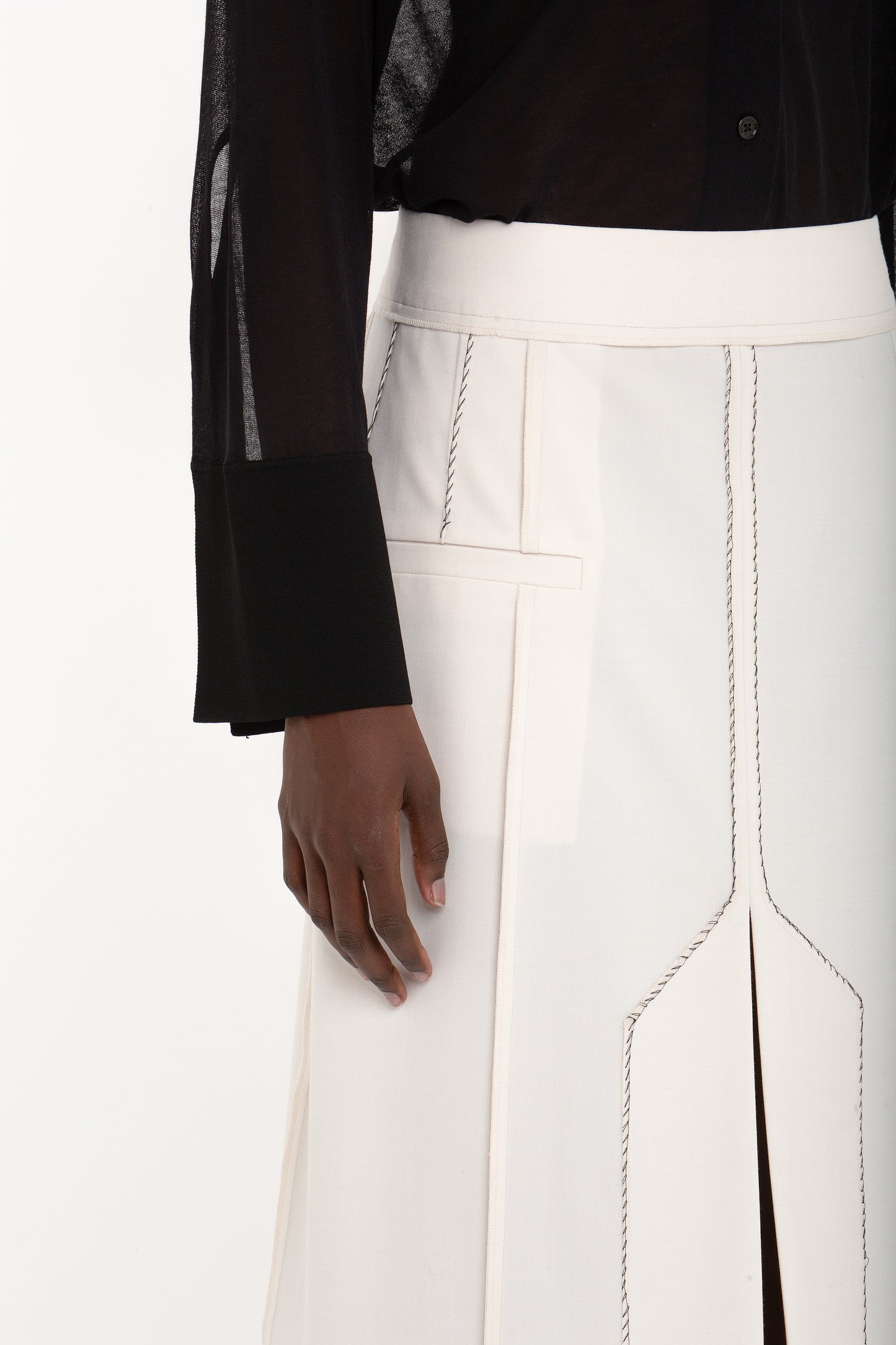 A person wearing a Victoria Beckham Pocket Detail Shirt In Black and a high-waisted white skirt with visible seams and a front slit. The person's right hand is resting by their side.