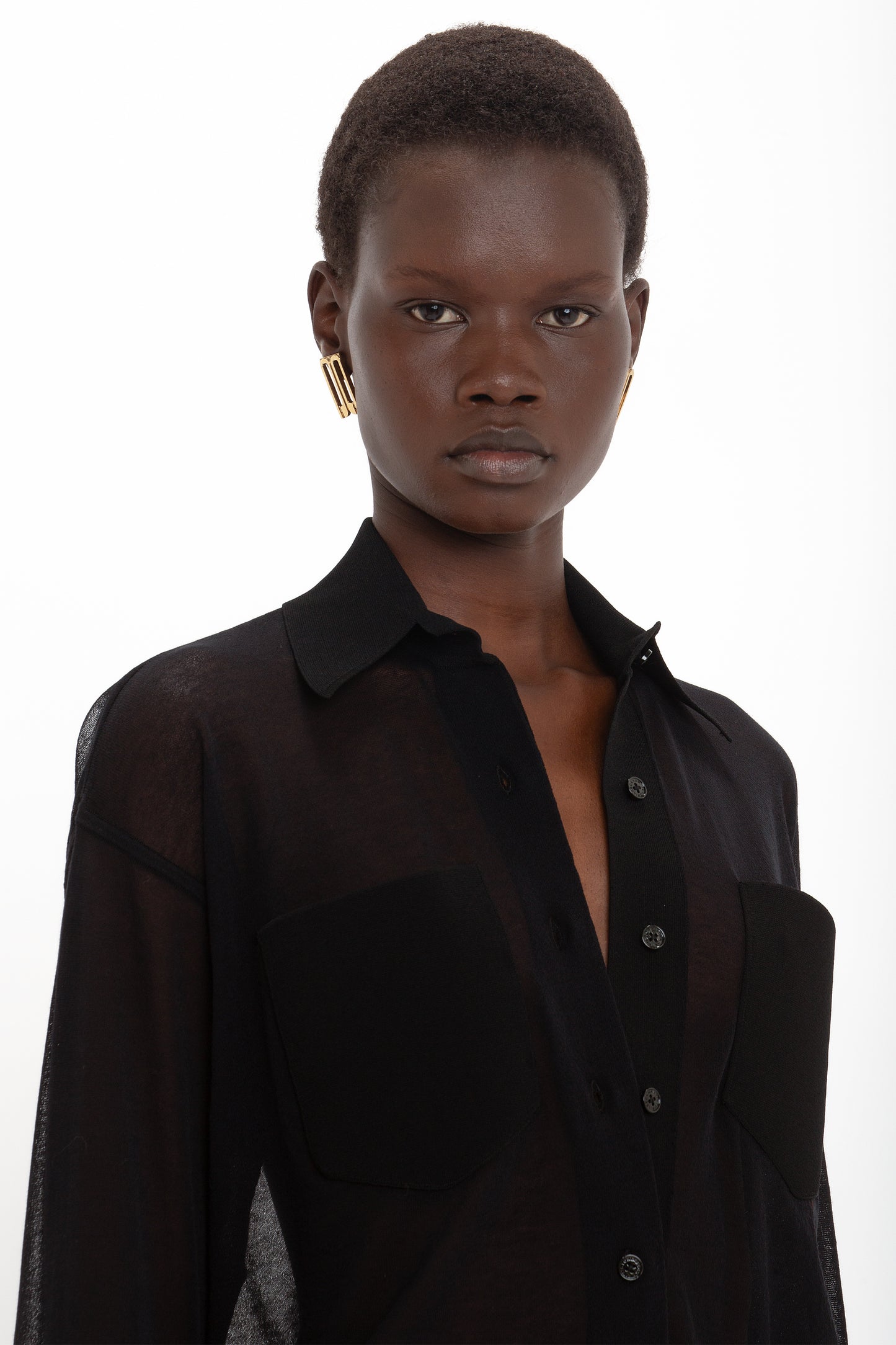 Person wearing a sheer black, relaxed-fit Pocket Detail Shirt In Black by Victoria Beckham with large front pockets and gold earrings, standing against a white background.