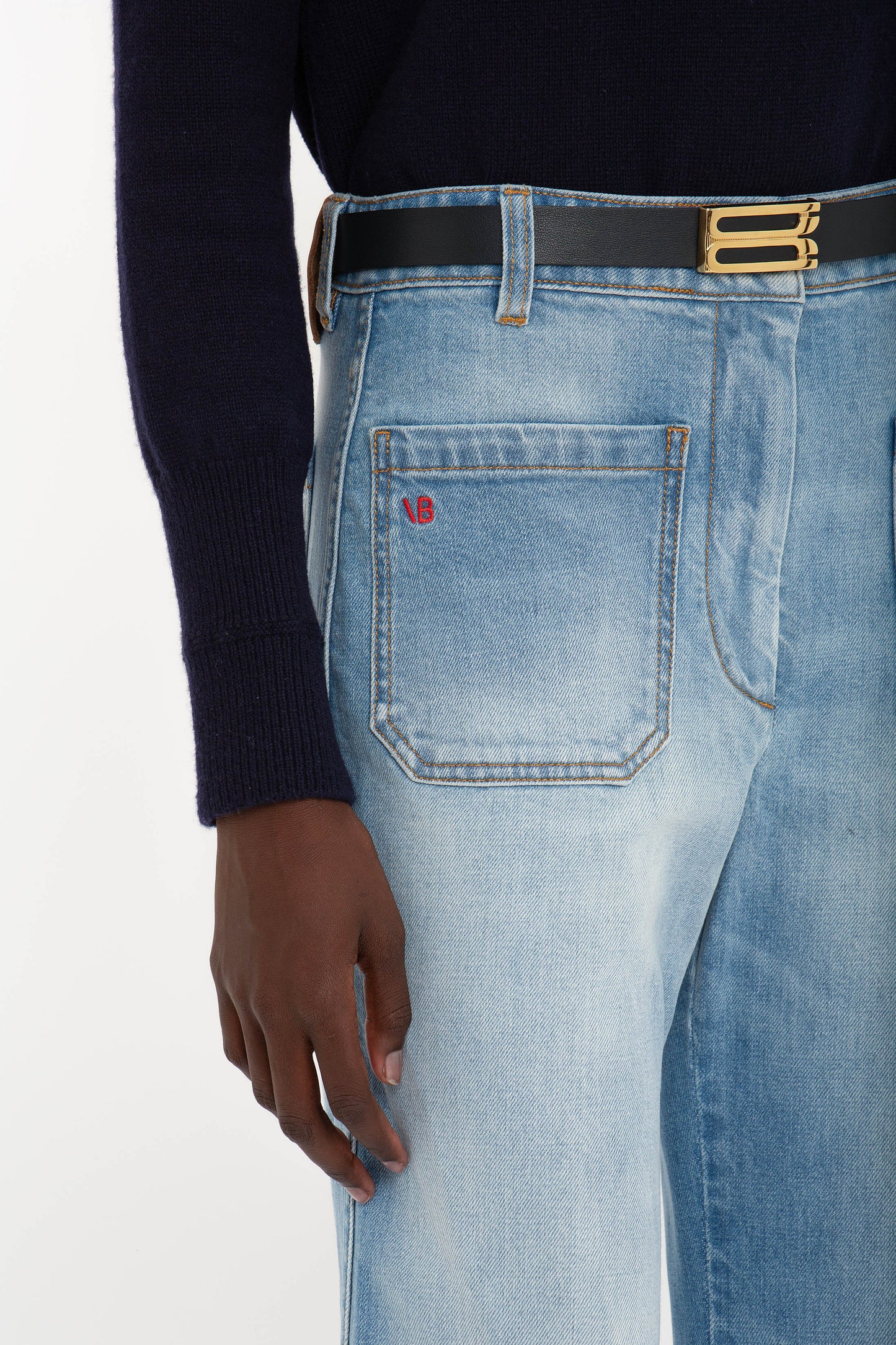 Person wearing Victoria Beckham Alina High Waisted Jean In Light Summer Wash in a vintage denim style with a red logo on the back pocket, a black belt with a gold buckle, and a dark long-sleeve top.