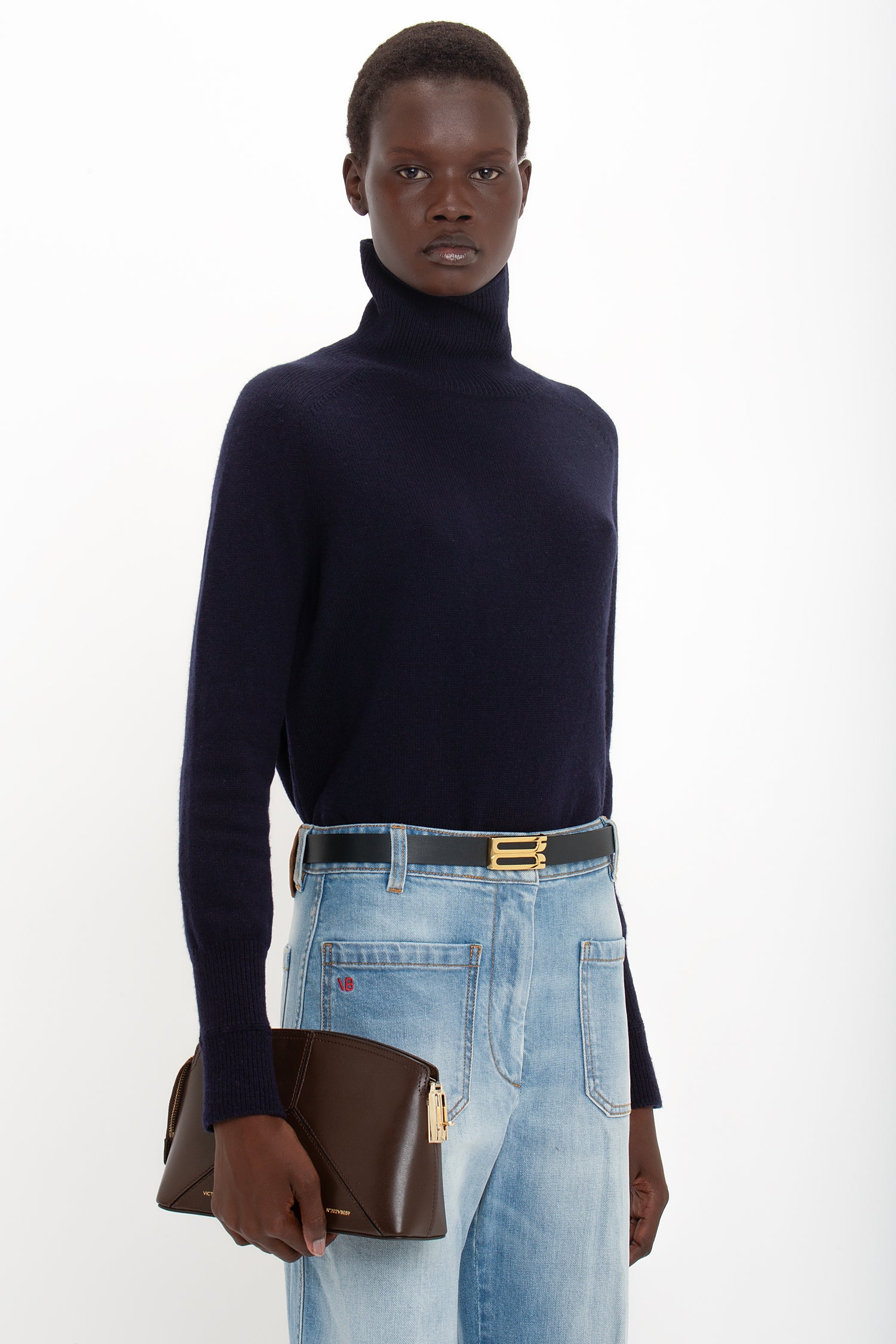 Person wearing a navy turtleneck sweater and Victoria Beckham Alina High Waisted Jean In Light Summer Wash, holding a brown clutch purse, standing against a white background.