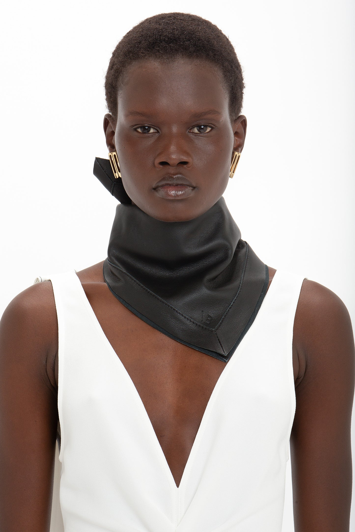 Person with short hair wearing a white sleeveless top, a classic Victoria Beckham Leather Scarf In Black, and gold geometric earrings against a white background.