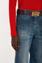 A person wearing a red sweater and blue jeans with a brown belt featuring a large gold buckle. The sweater's sleeve ends mid-hand, and a small red logo appears on the front pocket of the tailored Victoria Beckham Alina High Waisted Jean In Heavy Vintage Indigo Wash.