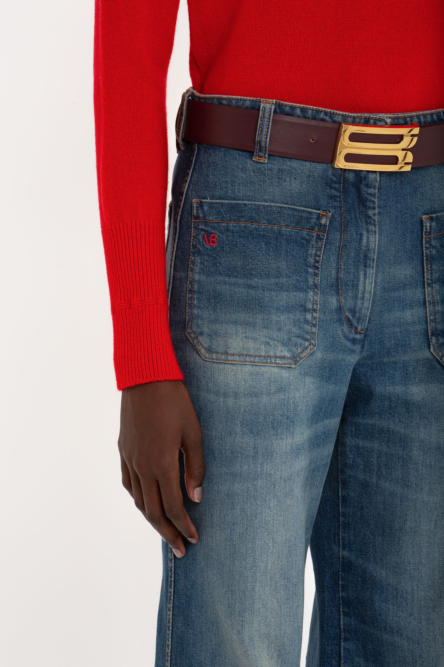 Person wearing a Victoria Beckham Polo Neck Jumper In Red, belted blue jeans with visible pocket stitching, and a brown belt with a gold buckle.