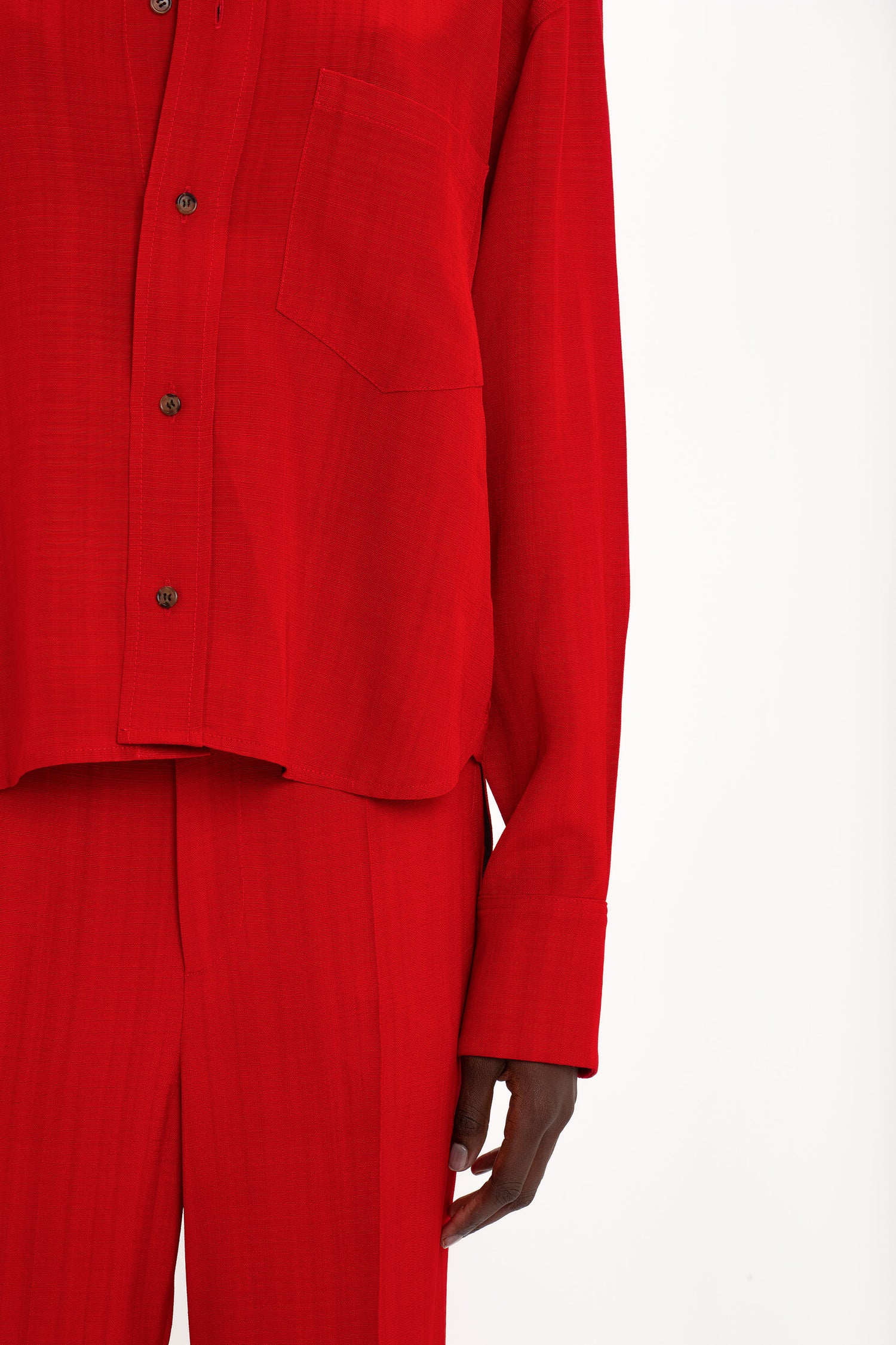 A person wearing a red masculine-inspired button-up Cropped Long Sleeve Shirt In Carmine by Victoria Beckham with a front pocket and matching red pants. The waist-defining cropped length adds a touch of elegance, while only the torso and upper legs are visible in the image.