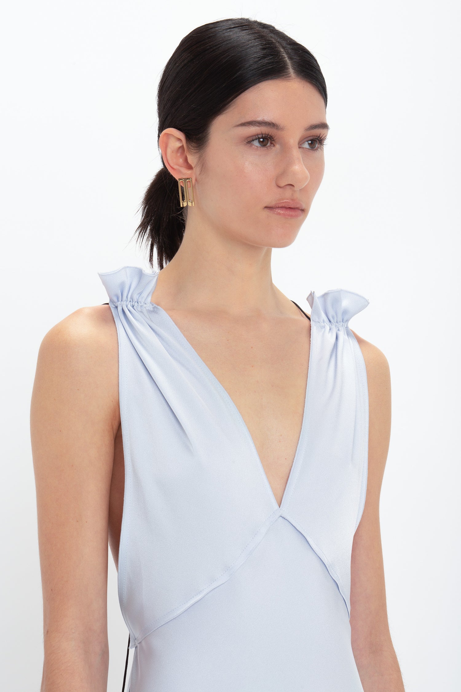 A woman with dark hair in a sleek, low ponytail models a pale blue Exclusive Gathered Shoulder Cami Floor-Length Gown In Ice Blue from Victoria Beckham crafted from crepe back satin. The sleeveless dress features a deep-V neckline and sophisticated gathered shoulder details. She pairs it elegantly with a geometric earring.