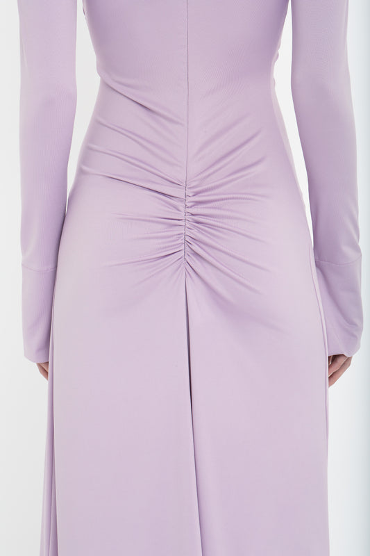 A close-up of the back of a person wearing a light purple Victoria Beckham Ruched Detail Floor-Length Gown In Petunia with long sleeves, crafted from stretch jersey and featuring a ruched detail at the center of the lower back.