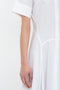 Close-up of a person's left arm and side, wearing a white short-sleeved, button-up Panelled Shirt Dress In White from Victoria Beckham made from organic cotton poplin. The background is plain white.