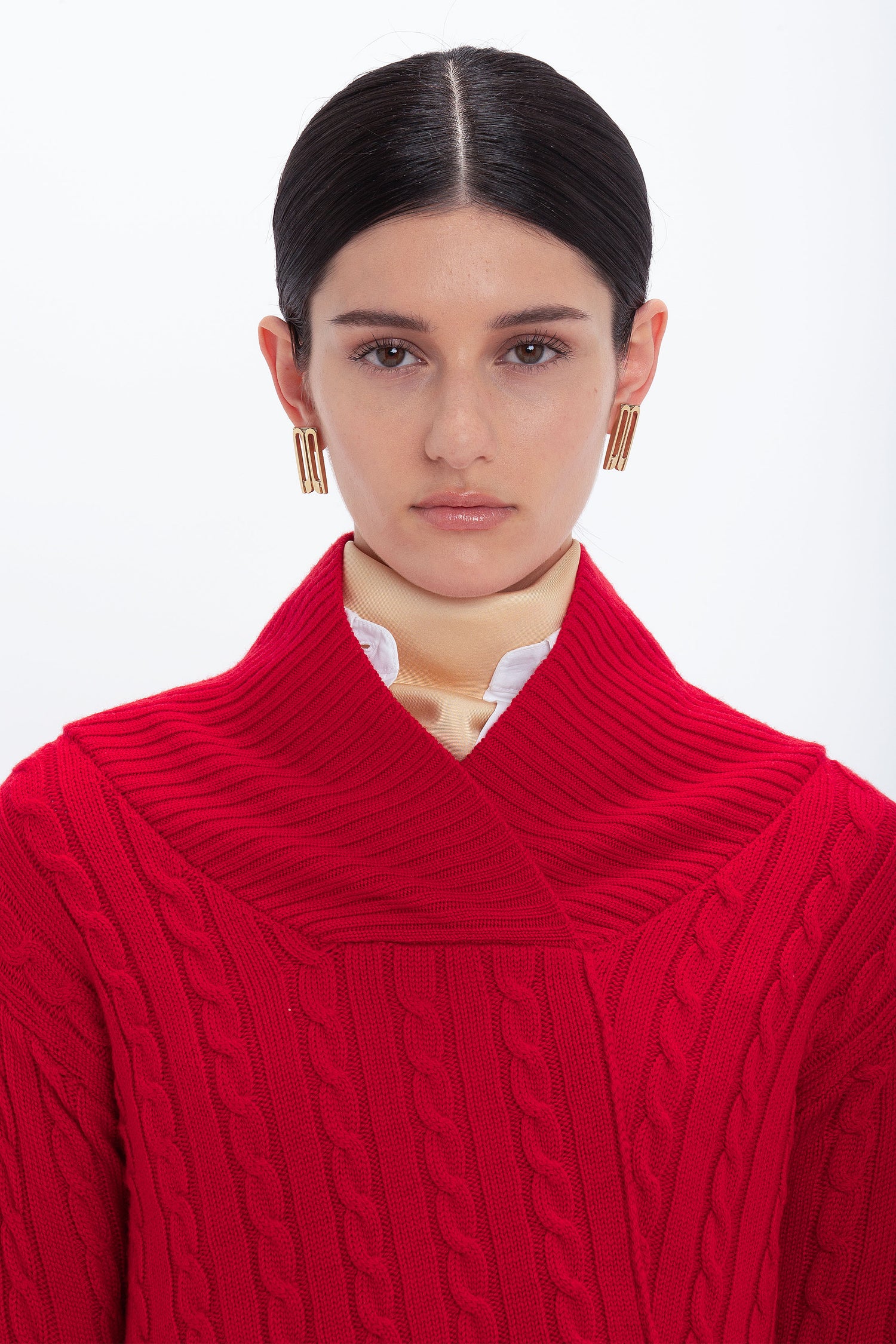 A woman with dark hair is wearing a red Wrap Detail Jumper In Red by Victoria Beckham and gold rectangular earrings, standing in front of a white background.