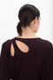 A person with dark hair tied in a low ponytail is wearing the Victoria Beckham Twist Detail Jersey Top In Deep Mahogany with a cutout design on the back, standing against a white background.